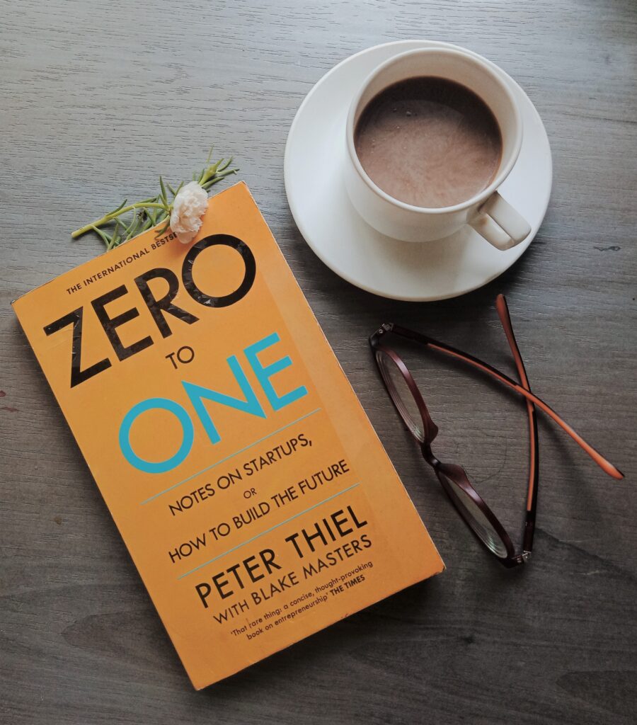 Zero to One book by Peter Thiel with Blake Masters is kept with specs and a cup of coffee.