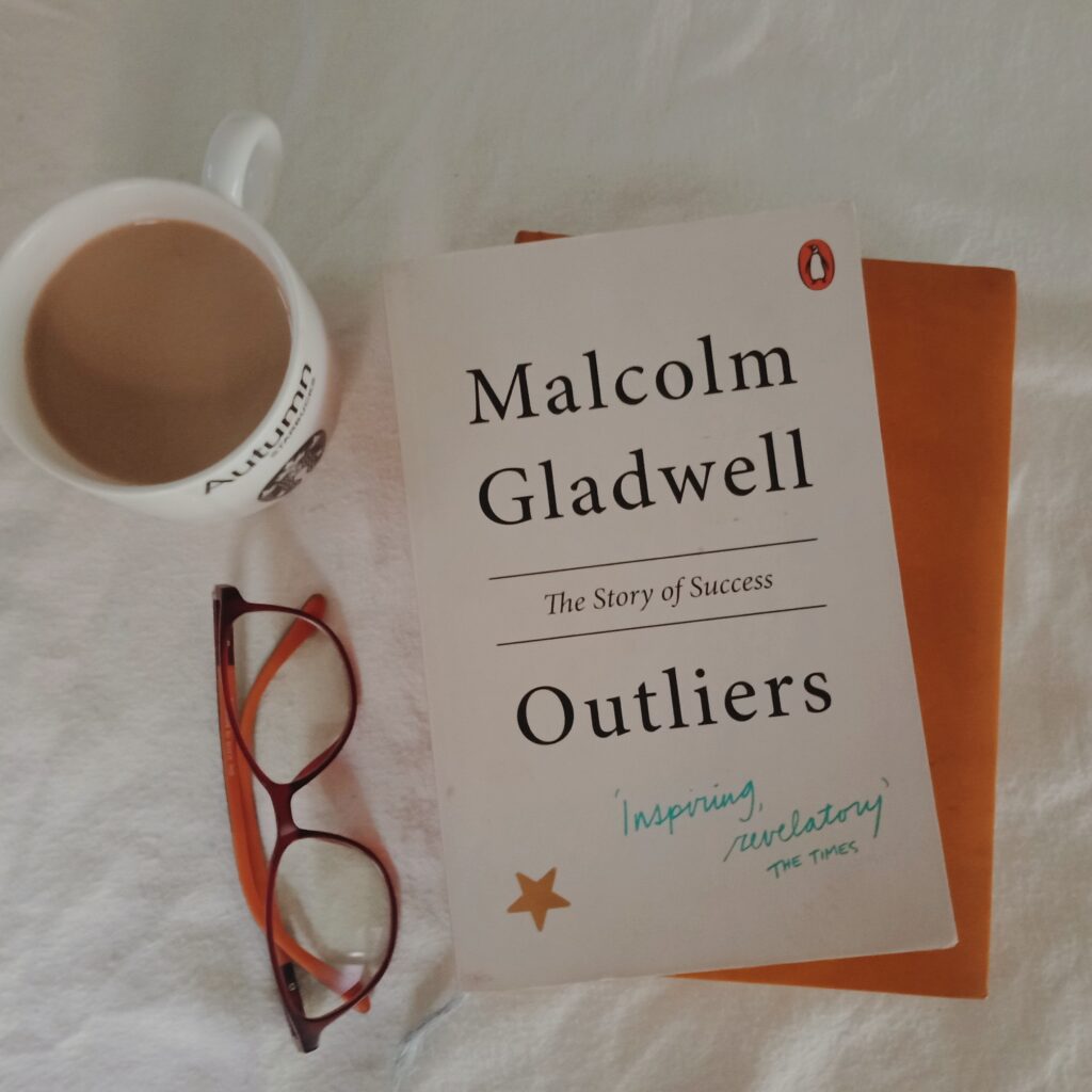 Book Outliers:The story of success by Malcolm Gladwell is kept with a cup of coffee and specs 